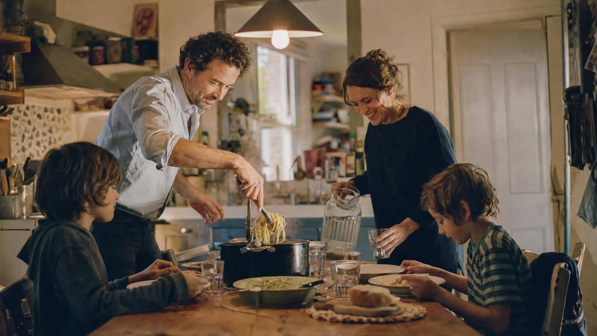 A father serving pasta to his children at the kitchen table in the evening