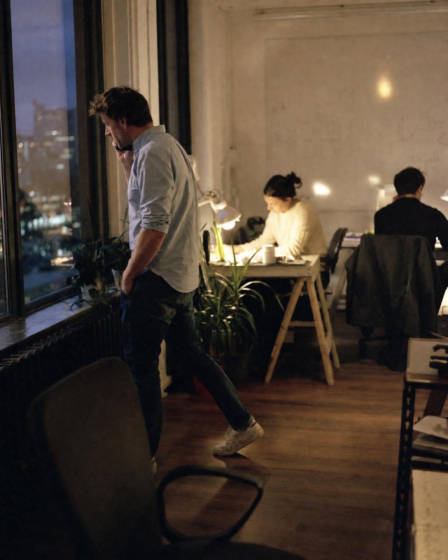 Ein Founder läuft bei einem späten Anruf am Fenster auf und ab, während sein Team unter warmen Lampen arbeitet